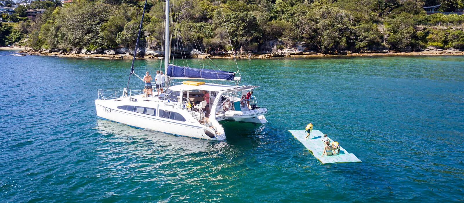 Pearl Catamaran anchored near a green shoreline with people on a raft.
