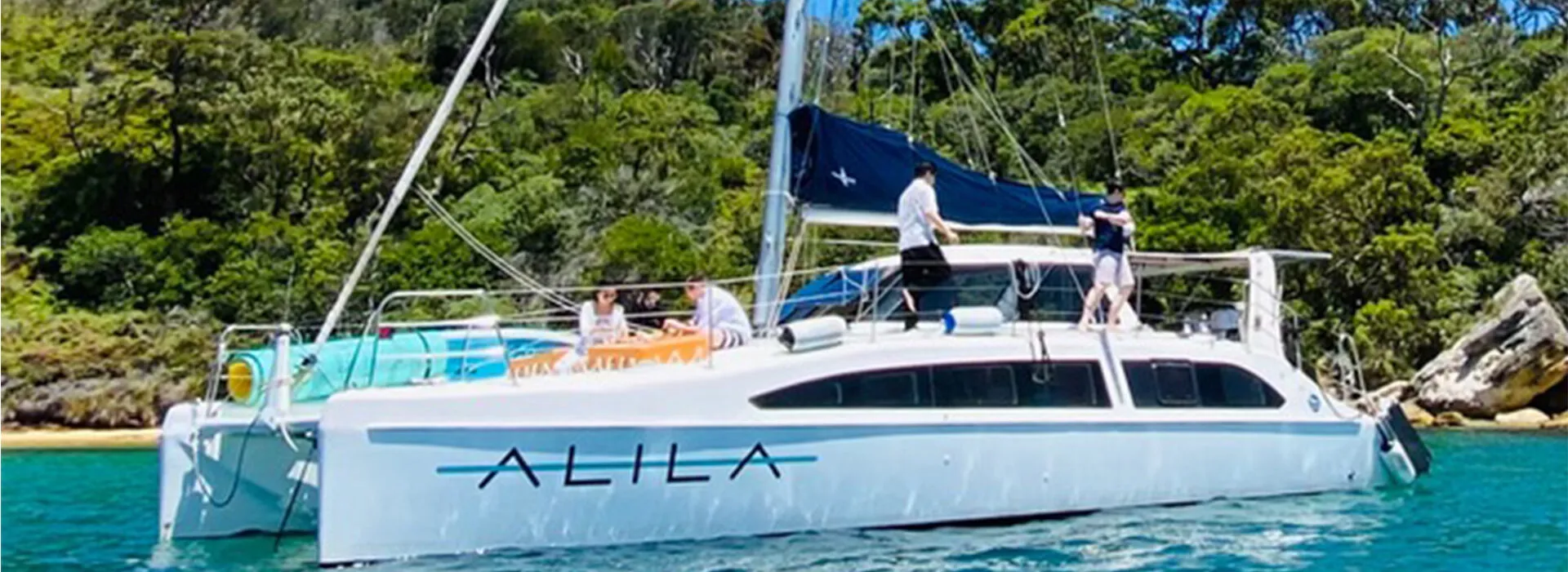 Alila yacht anchored near a lush green shoreline with people onboard. Sailing on the Alila yacht with guests enjoying the scenic surroundings.