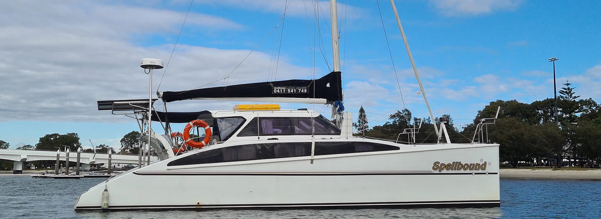 Sailboat named Spellbound docked on calm waters under a cloudy sky. Variation #1: Catamaran named Spellbound moored in tranquil waters with trees in background.